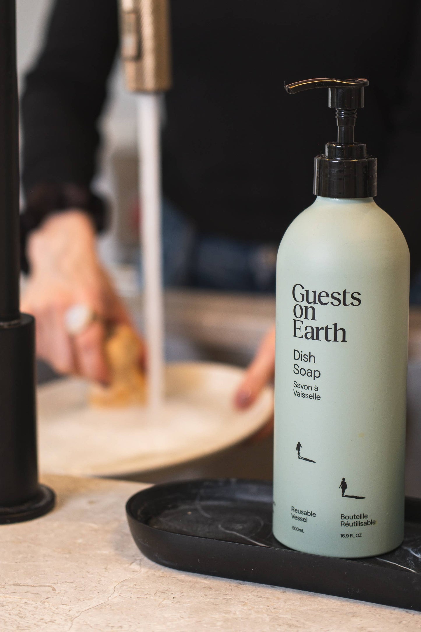Bottle of 'Guests on Earth' dish soap on a kitchen counter with a person washing dishes in the background.