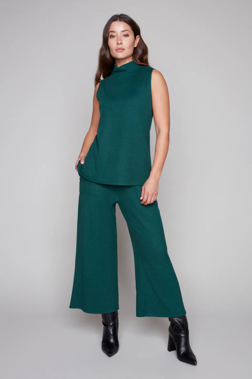 A woman wearing the Penelope Wide Leg Knit Pants by Compli K in Forest Green, cropped wide leg pants in thick textured knit with large patch opckets and an elastic waist. She is wearing them with the matching Pascale Mock Neck Tank and standing in front of a grey background.