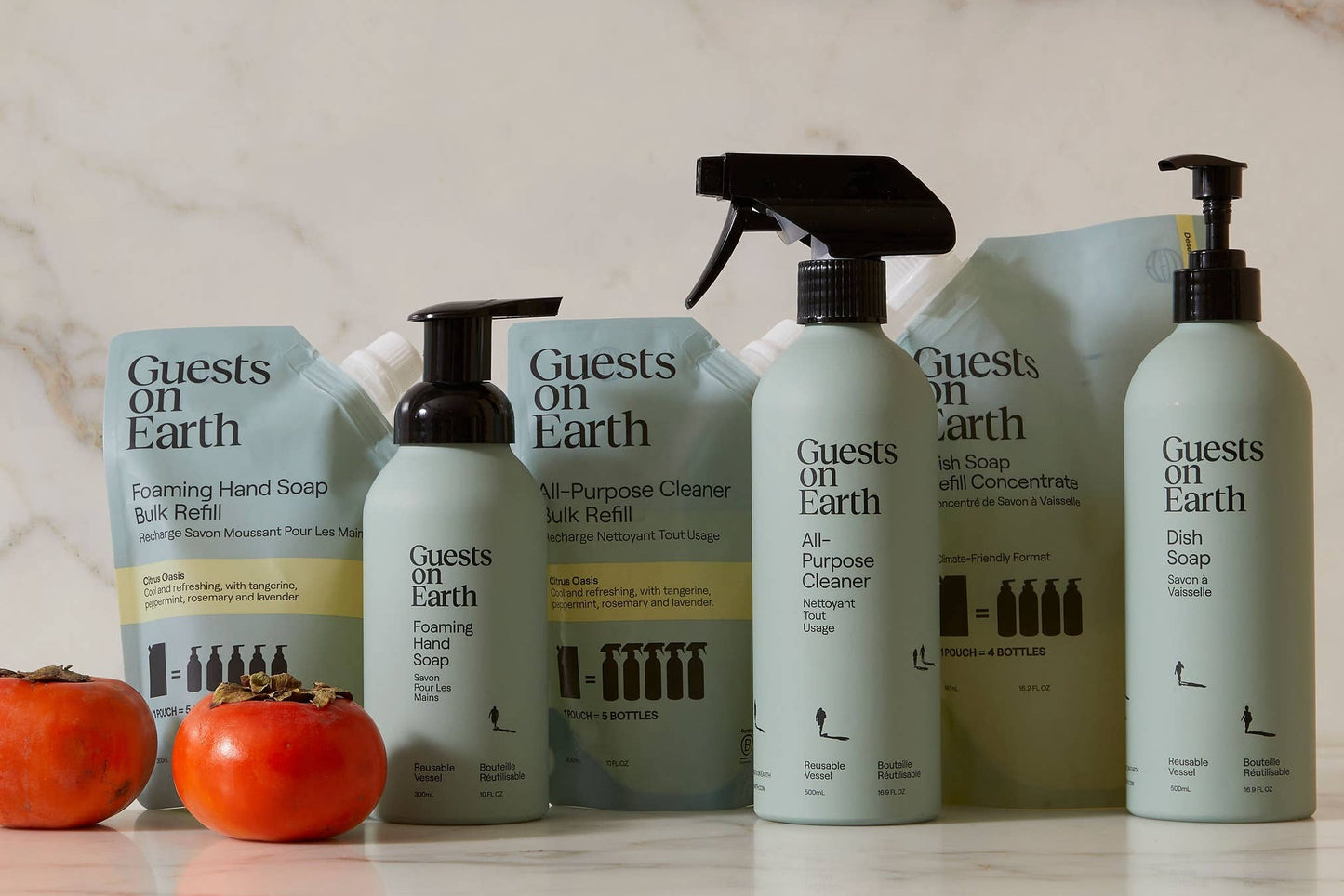 Set of 'Guests on Earth' cleaning products on a countertop with two persimmons.