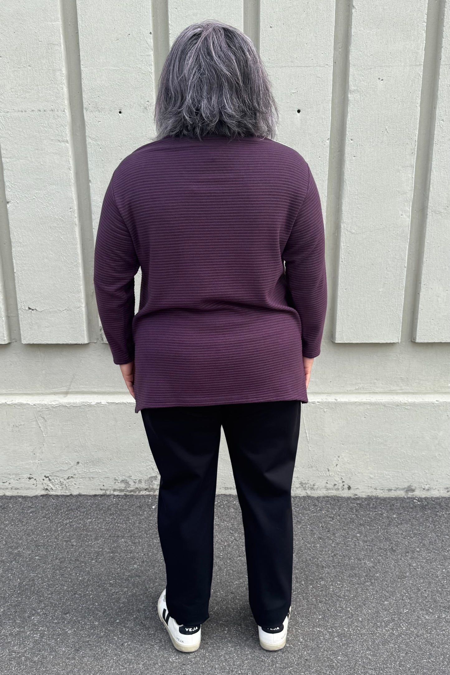 Back view of a woman wearing the Gladis Sweater by Solomia in Burgundy Pink Navy, a colour blocked in a mix of textures with a round neck, long sleeves, below the hip length ad side slits. She is wearing it with black pants and standing outside in front of a grey wall.