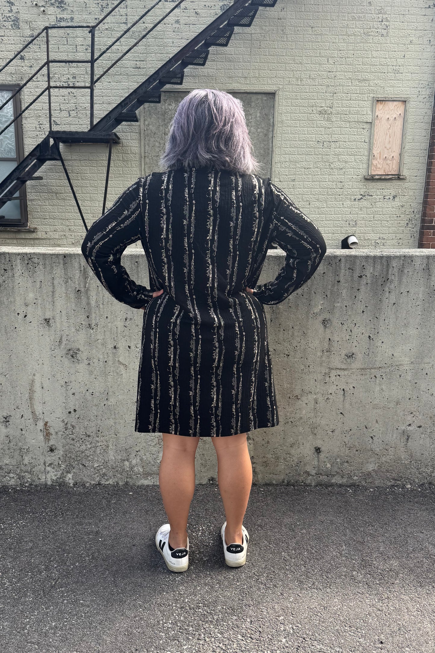 Back view of a woman wearing the Floella Dress by Pure Essence in Black/Tan, featuring a turtleneck, long sleeves, a slight a-line shape that hits the knee, and front patch pockets. She is standing in front of a grey wall.