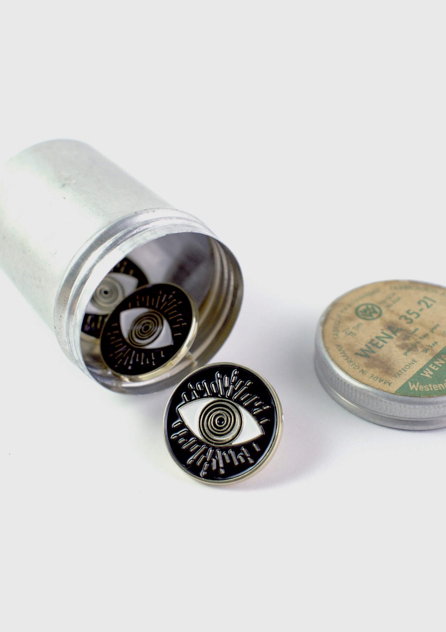 A number of The Eyeball pins by Badaboöm Studio, circular black enamel pins with a gold-tone metal border and an eye design in the centre, are shown spilling out of a silver container against a white background.