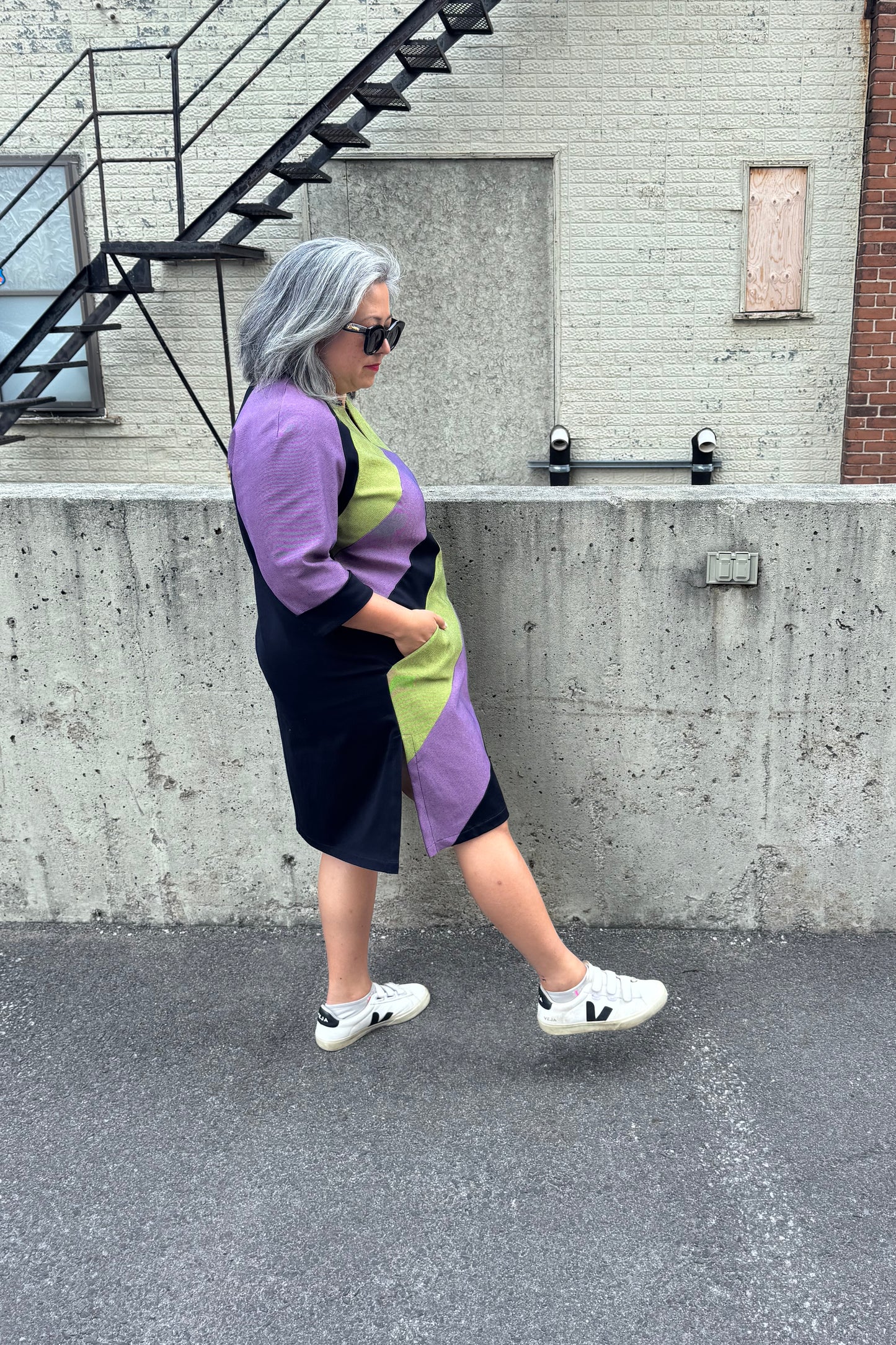 Side view of a woman wearing the Lorraine Dress by Solomia in Avocado/Purple Navy, an A-line colour-blocked dress in stretch denim with 3/4 sleeves and a knee-length hem. She is standing outside in front of a cement wall.