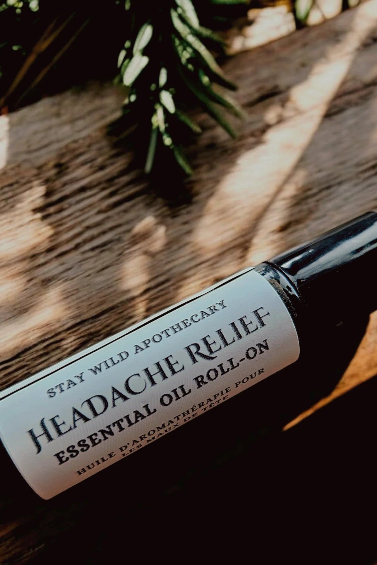 A bottle of 'Headache Relief' essential oil roll-on by Stay Wild Apothecary is shown on a wooden surface with greenery.