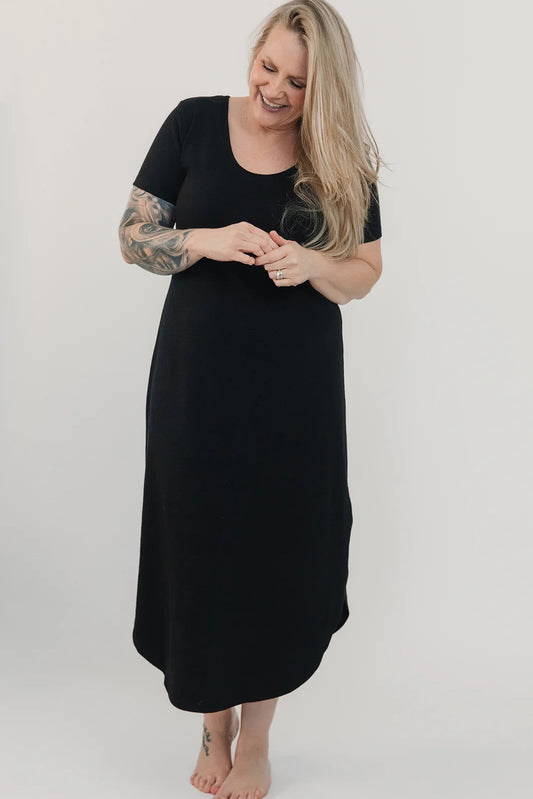 A woman wearing the Field Dress by Blondie in Black, with short sleeves, a scooped neck, and a contoured hem that hits at mid-calf and cuts up at the sides. She is standing in front of a white background. 