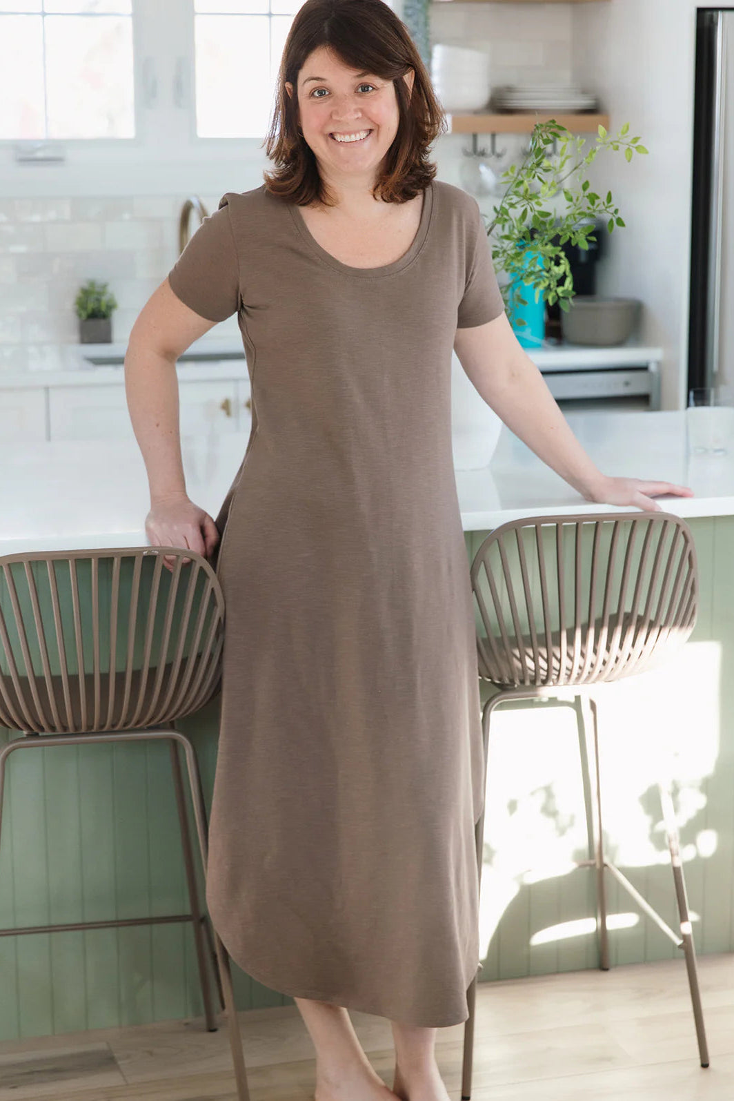 A woman wearing the Field Dress by Blondie in Earth, with short sleeves, a scooped neck, and a contoured hem that hits at mid-calf and cuts up at the sides. She is standing in a kitchen.