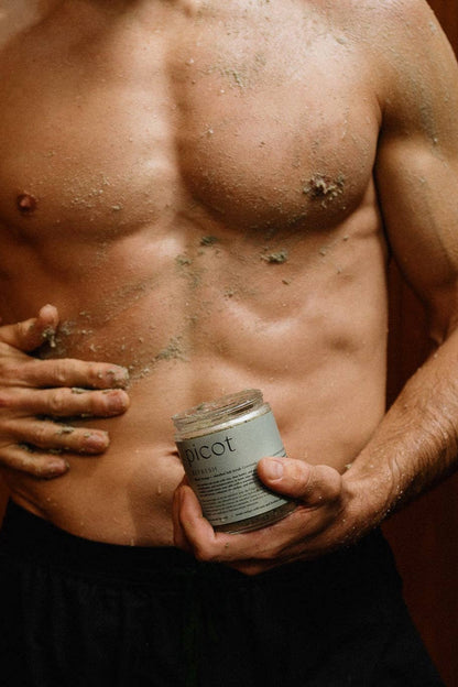 Man holding a container of Picot Refresh salt scrub and rubbing it into his torso with a brown background