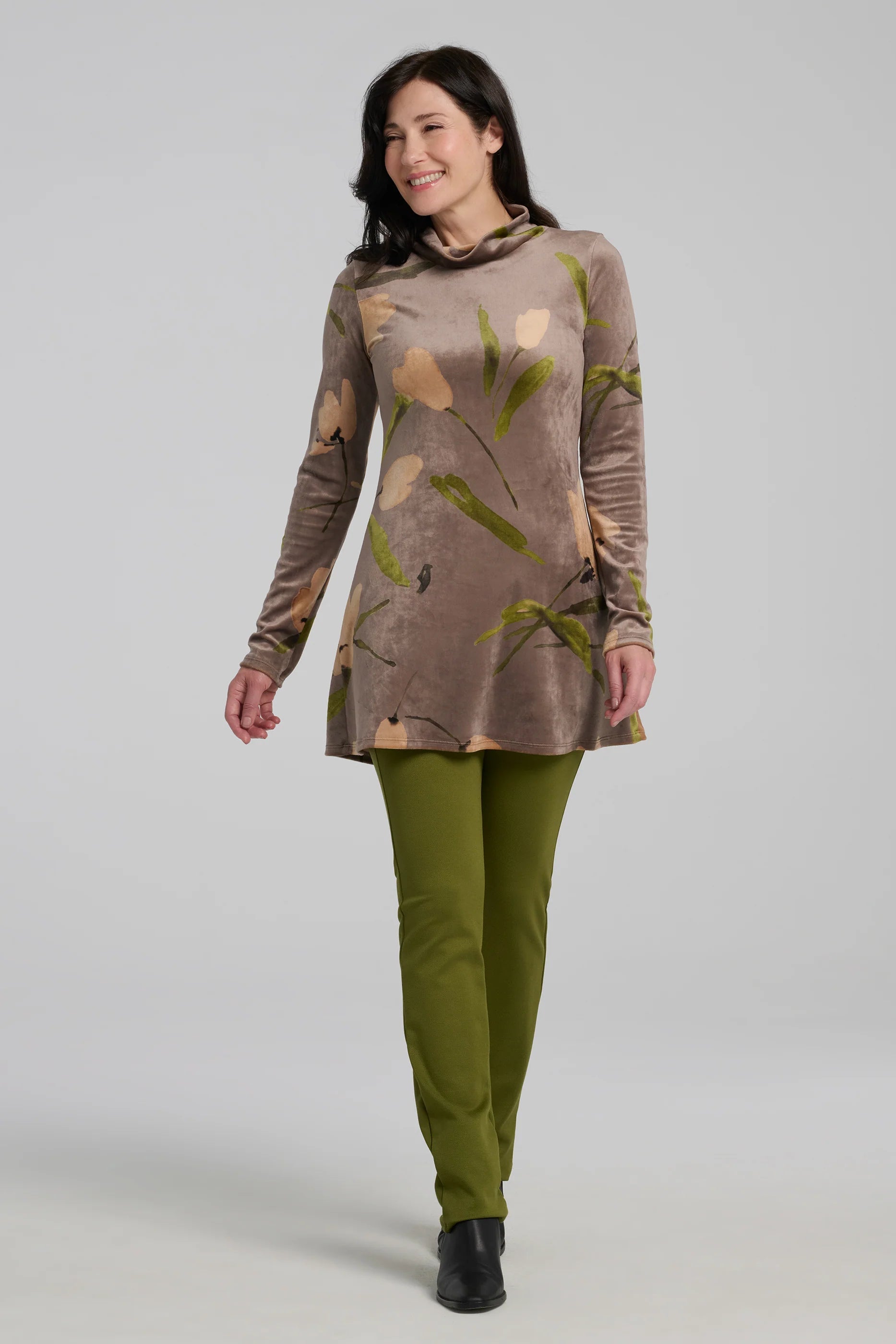 A woman wearing the Holly Tunic by Kollontai in Taupe, an Italian velvet A-line tunic with a turtleneck, long sleeves, and a floral print. She is wearing it with the Bari Pants in Green and standing in front of a grey background. 