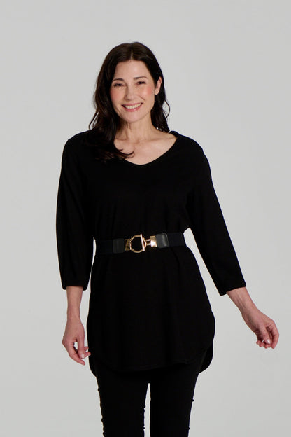  A woman wearing the Clara Tunic by Kollontai in Black, a stretch corduroy piece with a V-neck, a rounded hem, 3/4 sleeves, and inverted pleats at the shoulders. She is wearing it belted over black leggings and standing against a grey background. 