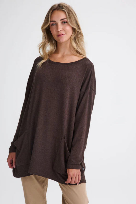 A woman wearing the Auclair Top by Message Factory in Brown, a bamboo/cotton/modal knit with a wide neck, long drop sleeves, a hi-low hem, and front patch pockets. She is wearing it with tan pants and standing in front of a white background. 