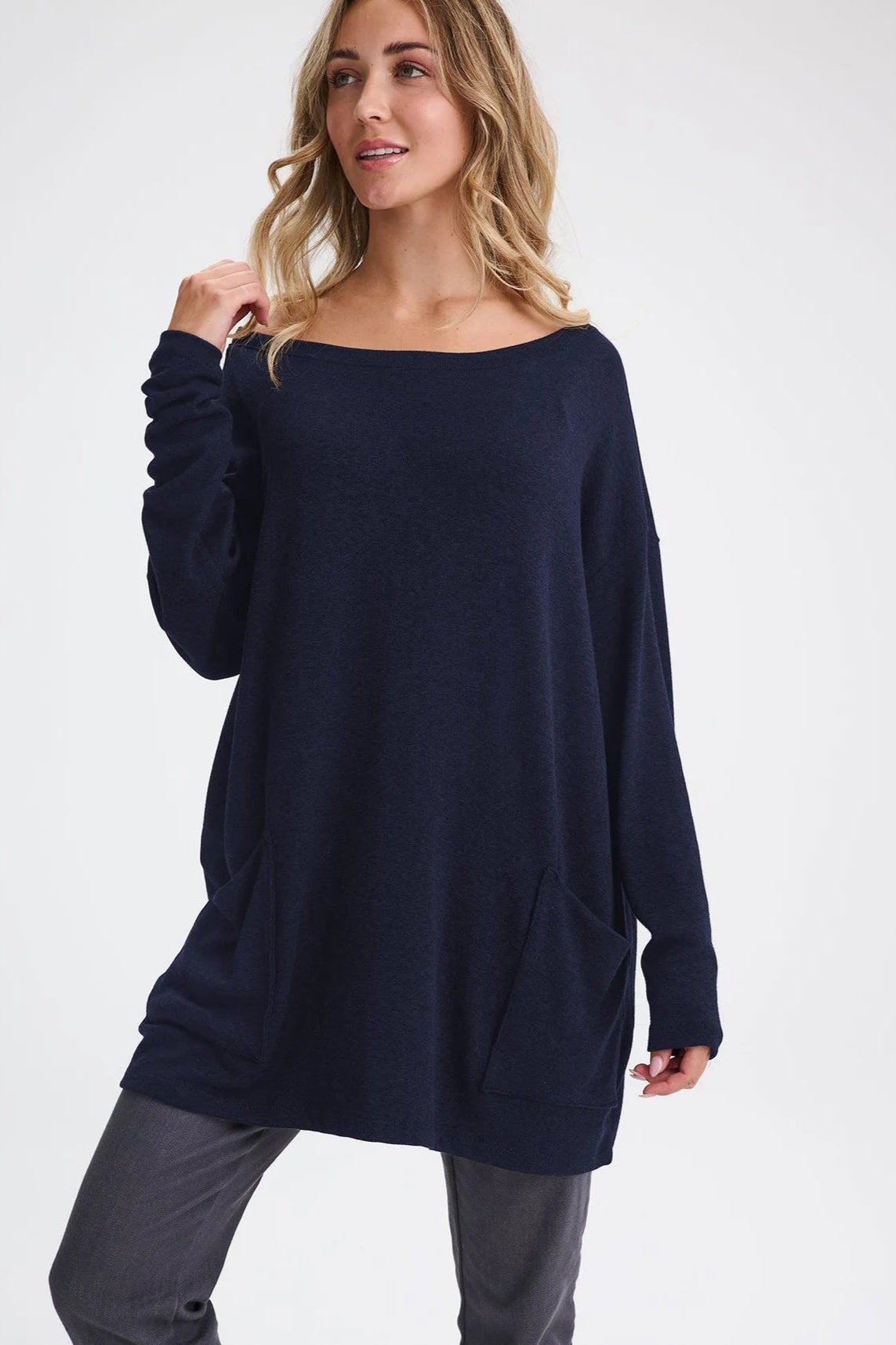 A woman wearing the Auclair Top by Message Factory in Navy, a bamboo/cotton/modal knit with a wide neck, long drop sleeves, a hi-low hem, and front patch pockets. She is wearing it with tan pants and standing in front of a white background. 