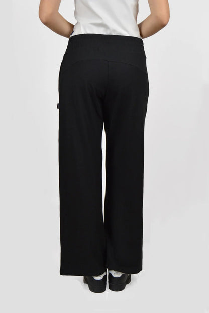 Back waist down view of the Tadoussac Pants by Message Factory in Black: flared sweatpants with an elastic waist in a bamboo/cotton blend. She is wearing a white tank with them and is standing in front of a white background. 