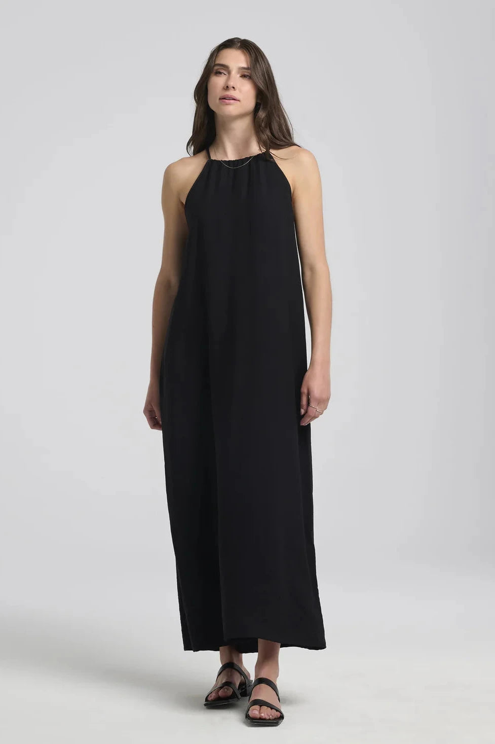 A woman wearing the Anwen Dress by Kollontai in Black, a loose-fitting rayon maxi dress with an adjustable halter neckline. She is standing in front of a white background. 