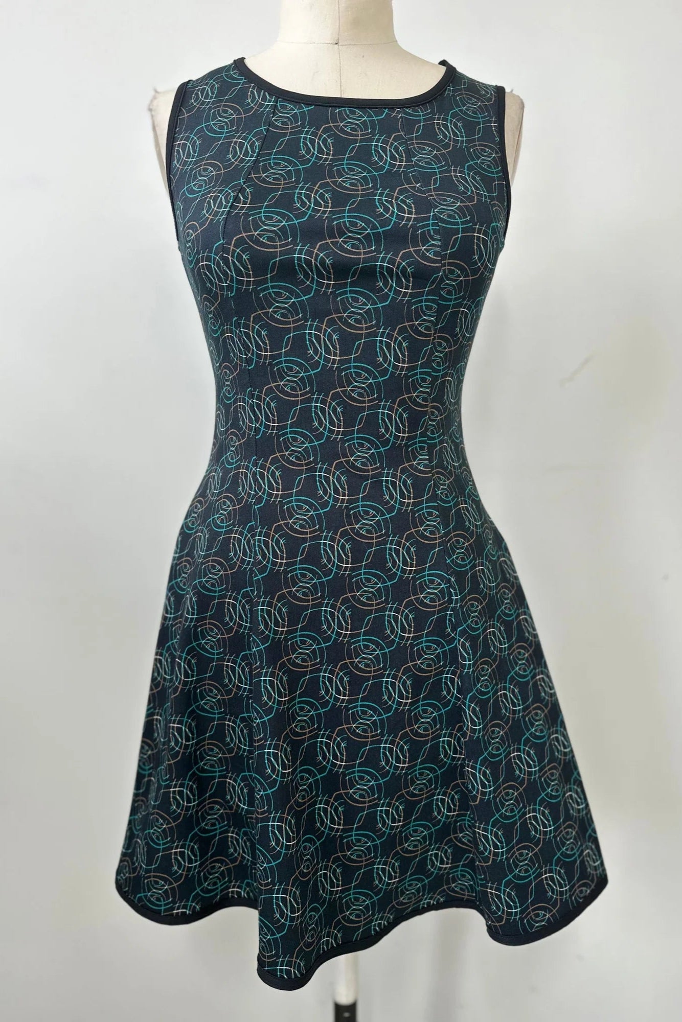 The Silba Dress by Kollontai in a Turquoise on black print, a sleeveless dress with a round neck, fit and flare shape, and above the knee hem, is shown on a mannequin against a white background. 