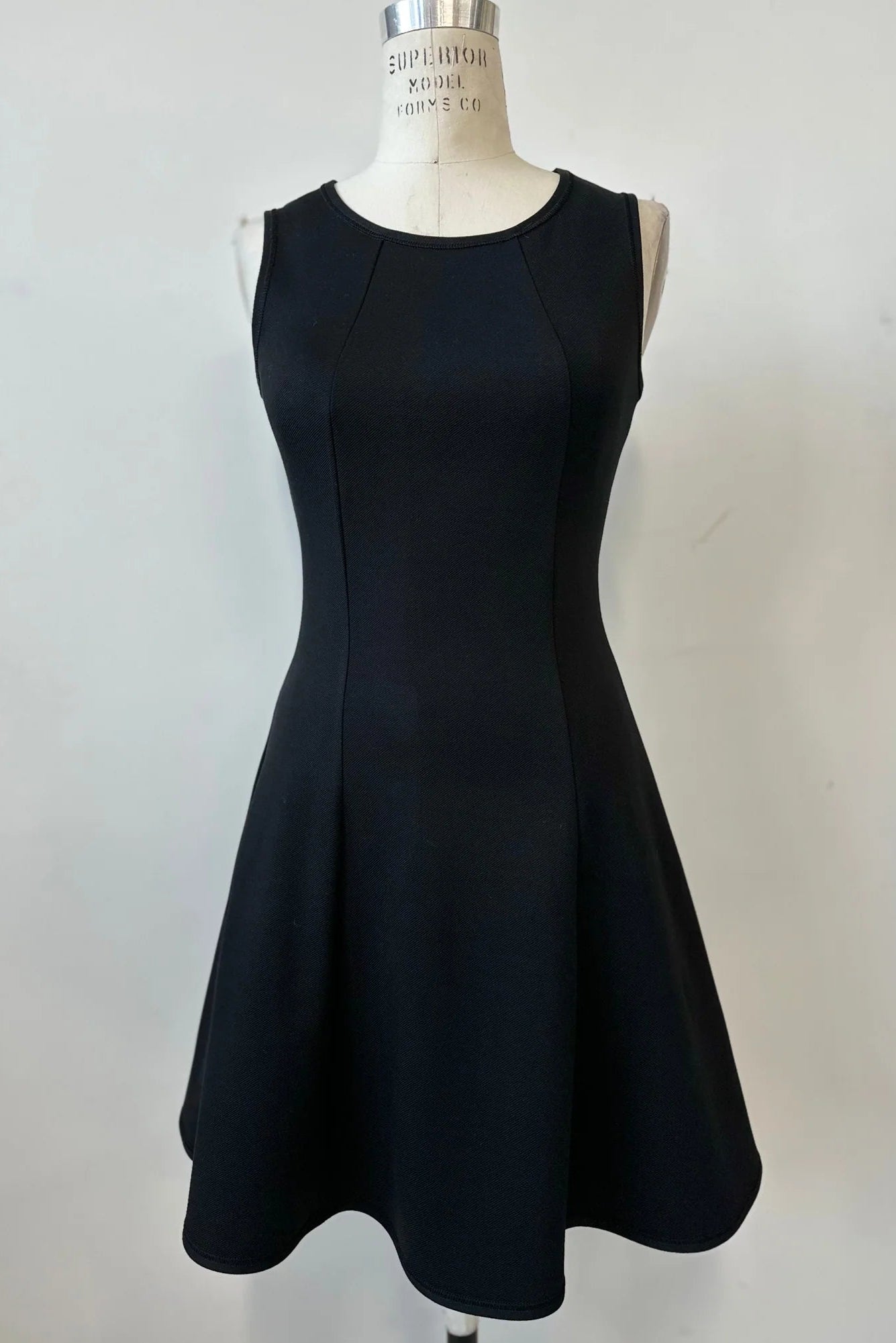 The Toscana Dress by Kollontai in Black, a sleeveless piece with a round neck, princess seams, and fit and flare shape with a full skirt that hits above the knee, is shown on a mannequin against a white background. 