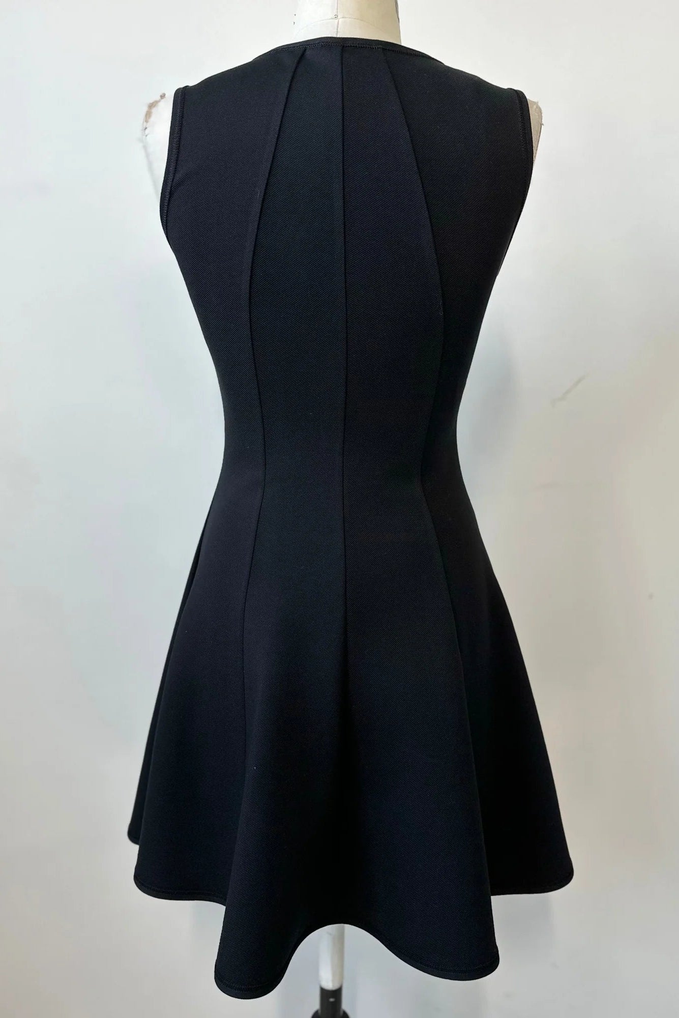 A back view of the Toscana Dress by Kollontai in Black, a sleeveless piece with a round neck, princess seams, and fit and flare shape with a full skirt that hits above the knee, is shown on a mannequin against a white background. 
