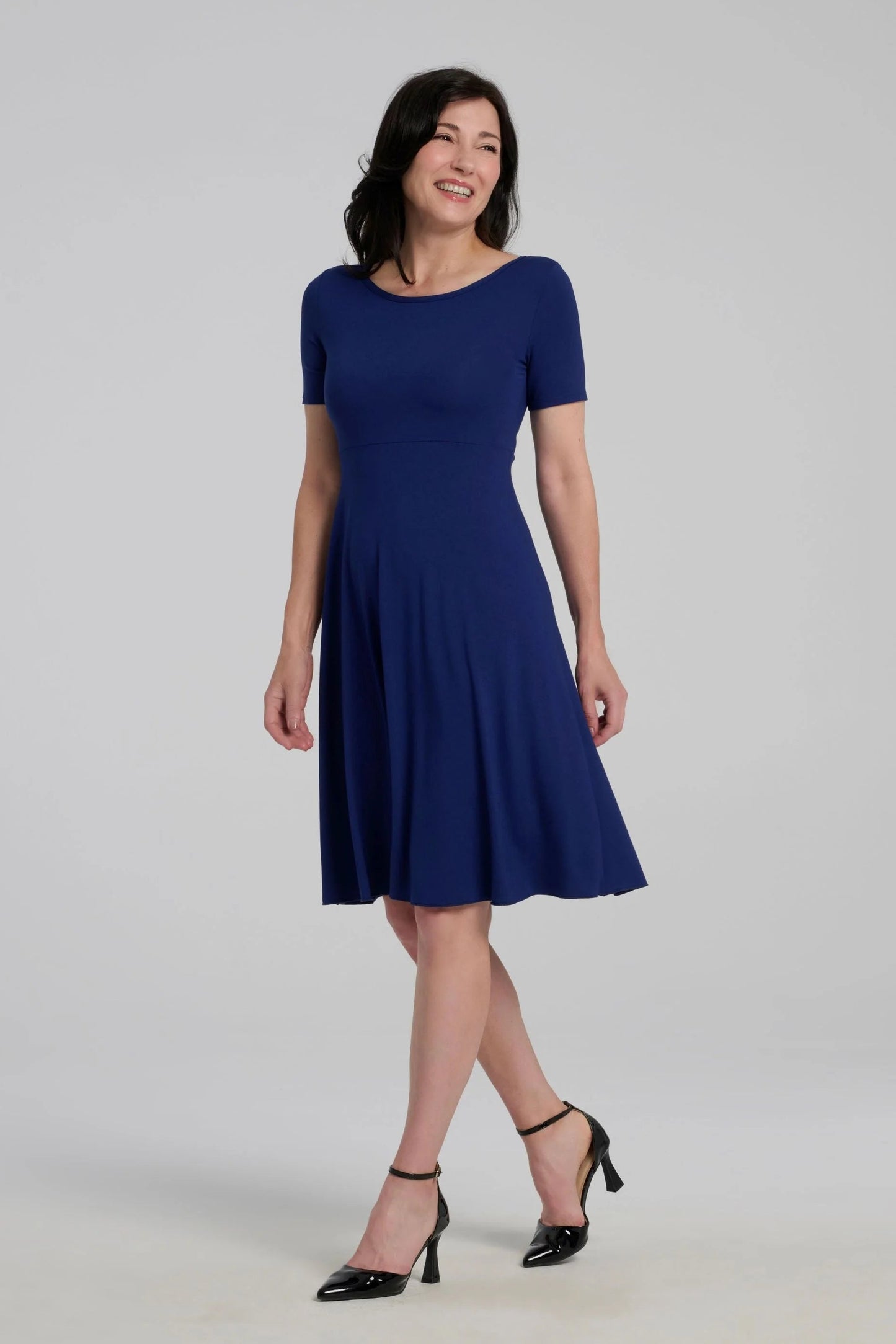 A woman wearing the Luma dress by Kollantai in Admiral blue, featuring short sleeves, a wide neck, an empire waist, and a full flared skirt that falls to the knee. She is standing in front of a grey background. 