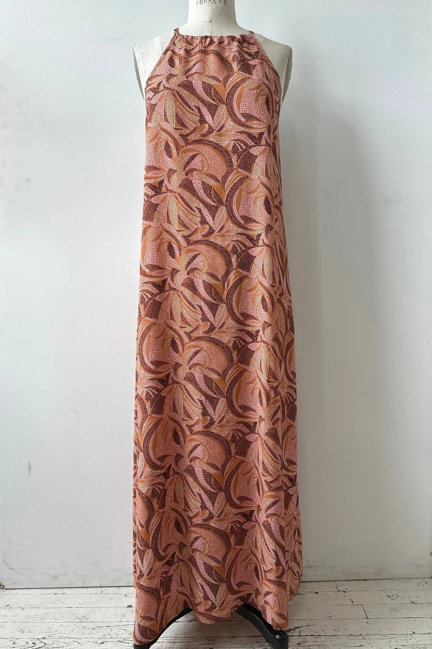 The Anwen Dress by Kollontai in Cinnamon print, a loose-fitting viscose maxi dress with an adjustable halter neckline, is show on a mannequin in front of a white background. 