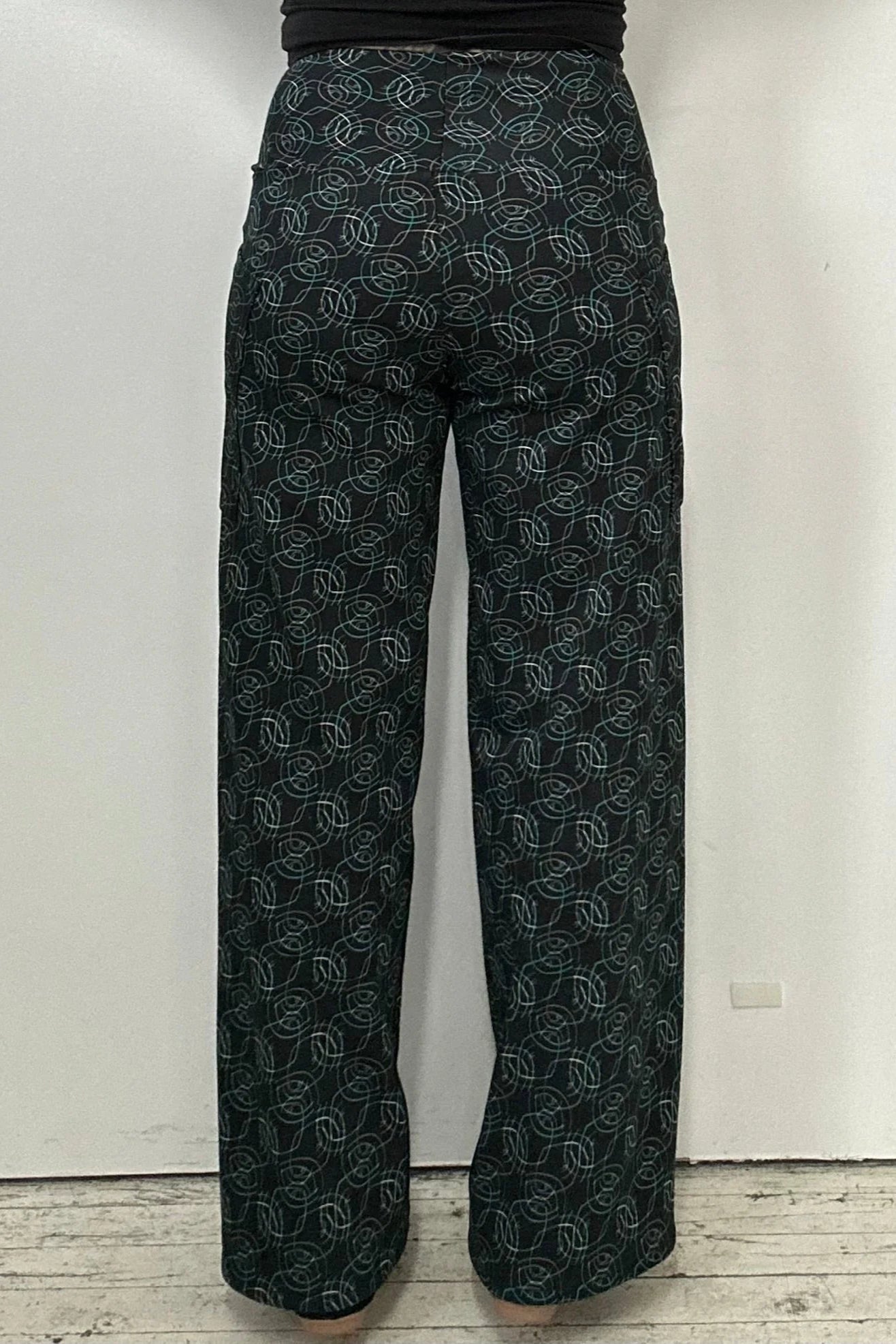 Back waist down view of a woman wearing the Elma Pants by Kollontai in Turquoise on black print, straight legged pants with a wide elastic waist and large slant pockets. 