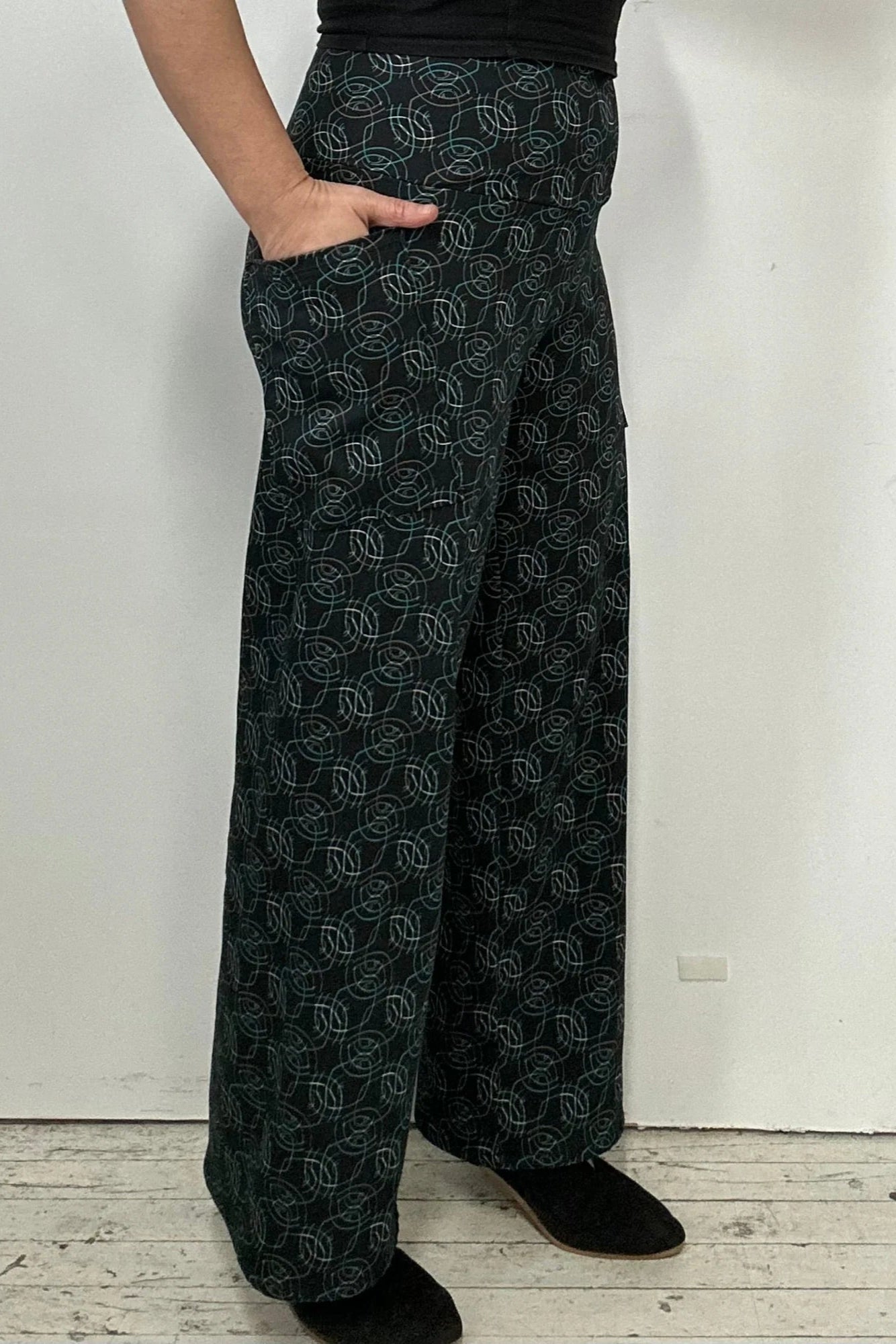 Waist down view of a woman wearing the Elma Pants by Kollontai in Turquoise on black print, straight legged pants with a wide elastic waist and large slant pockets. 