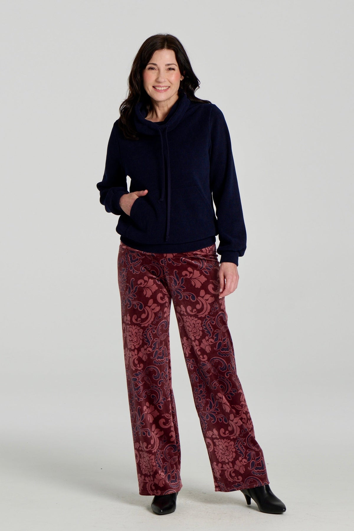 A woman wearing the Ramos Sweater by Kollontai with floral burgundy velvet pants. The Sweater is made from a navy ribbed fabric and has a large cowl neck with a drawstring and a kangaroo pocket. She is standing against a white background. 