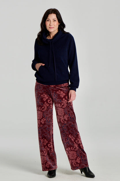 A woman wearing the Ramos Sweater by Kollontai with floral burgundy velvet pants. The Sweater is made from a navy ribbed fabric and has a large cowl neck with a drawstring and a kangaroo pocket. She is standing against a white background. 