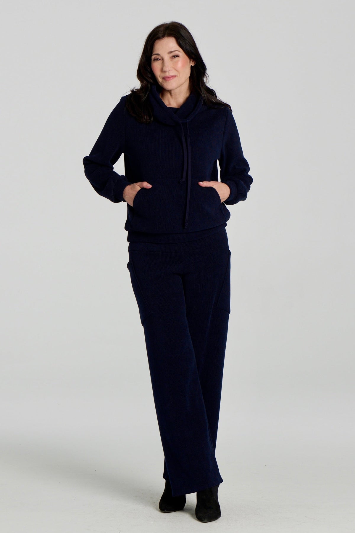 A woman wearing the Lorenzo Pants by Kollontai with the Ramos Sweater, both in a ribbed navy corduroy-like fabric. The Sweater has a large cowl neck with a drawstring and a kangaroo pocket, the pants have a wide pull on waist, straight legs, and large patch pockets. She is standing against a white background. 