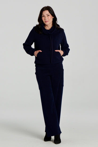 A woman wearing the Lorenzo Pants by Kollontai with the Ramos Sweater, both in a ribbed navy corduroy-like fabric. The Sweater has a large cowl neck with a drawstring and a kangaroo pocket, the pants have a wide pull on waist, straight legs, and large patch pockets. She is standing against a white background. 