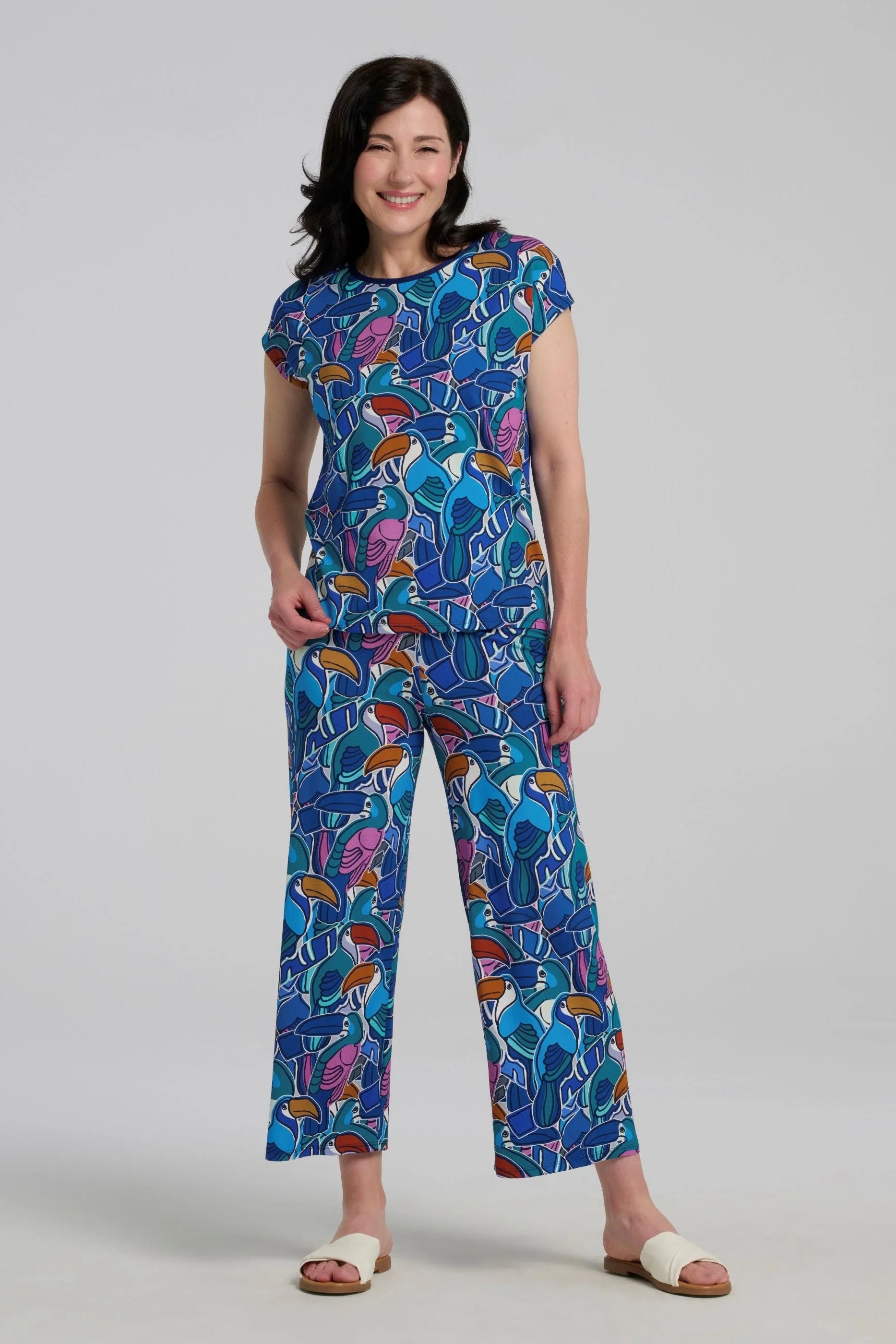 Woman wearing the Hanni Top by Kollontai in Admiral, a cap sleeved relaxed top with a jewel-toned parrot pattern on the front a solid blue back. She is wearing it with matching pants and standing in front of a grey background. 