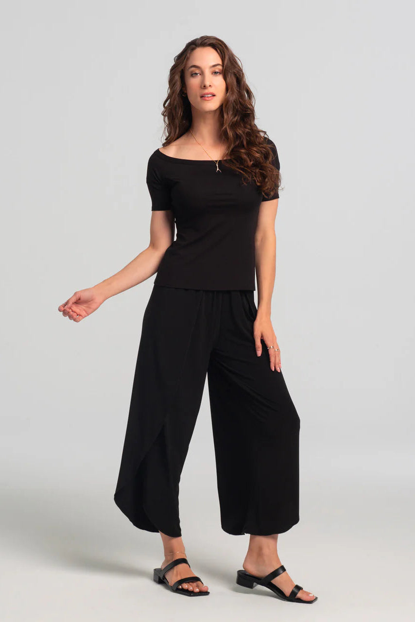 A woman wearing the Juno Top by Kollontai in Black, an off-the-shoulder top with short sleeves. She is wearing it with black harem pants against a grey background.