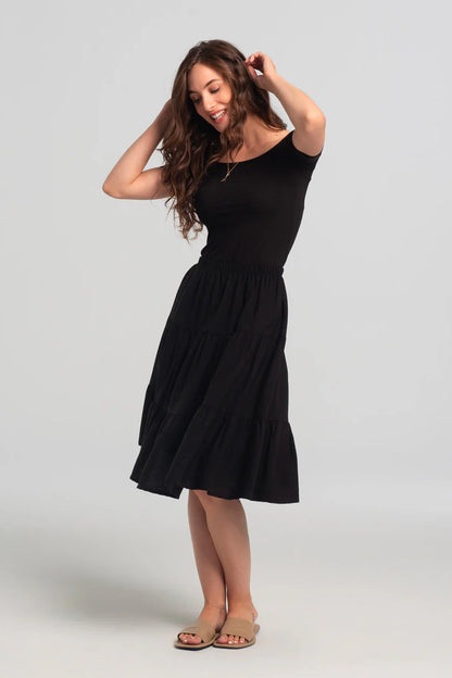 A woman wearing the Juno Top by Kollontai in Black, an off-the-shoulder top with short sleeves. She is wearing it with a black skirt against a grey background.