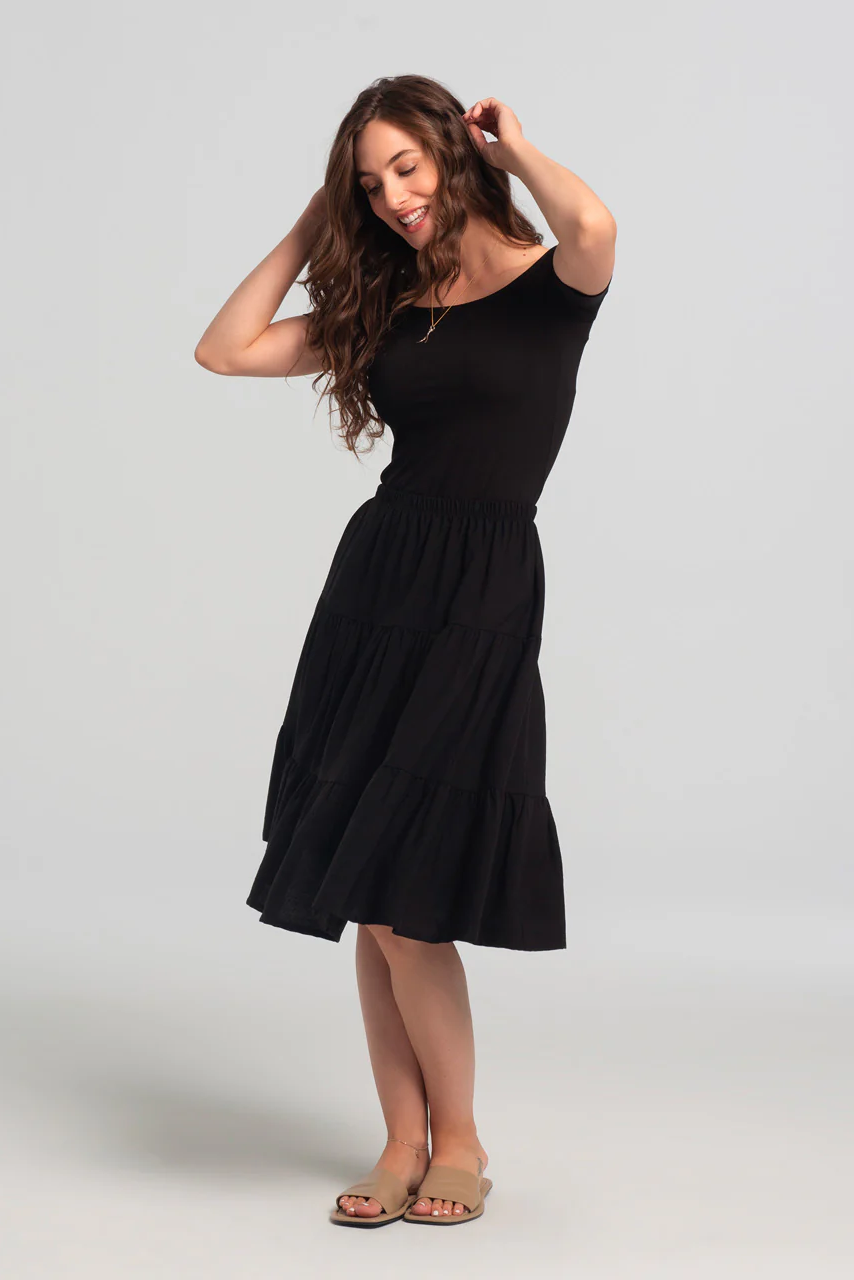 A woman wearing the Al-Juzza Skirt by Kollontai in Black with a black top, standing in front of a white background 