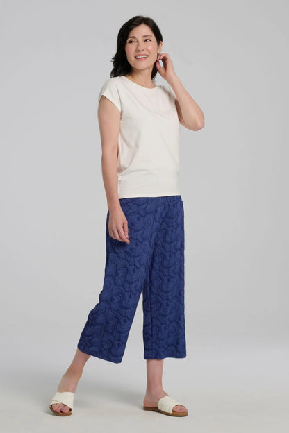 Woman wearing the Sana Pants by Kollonta in Atlantis Blue, slightly cropped straight leg pants with side patch pockets in a curlicue patterned jacquard fabric. She is wearing them with a white top against on a grey background.