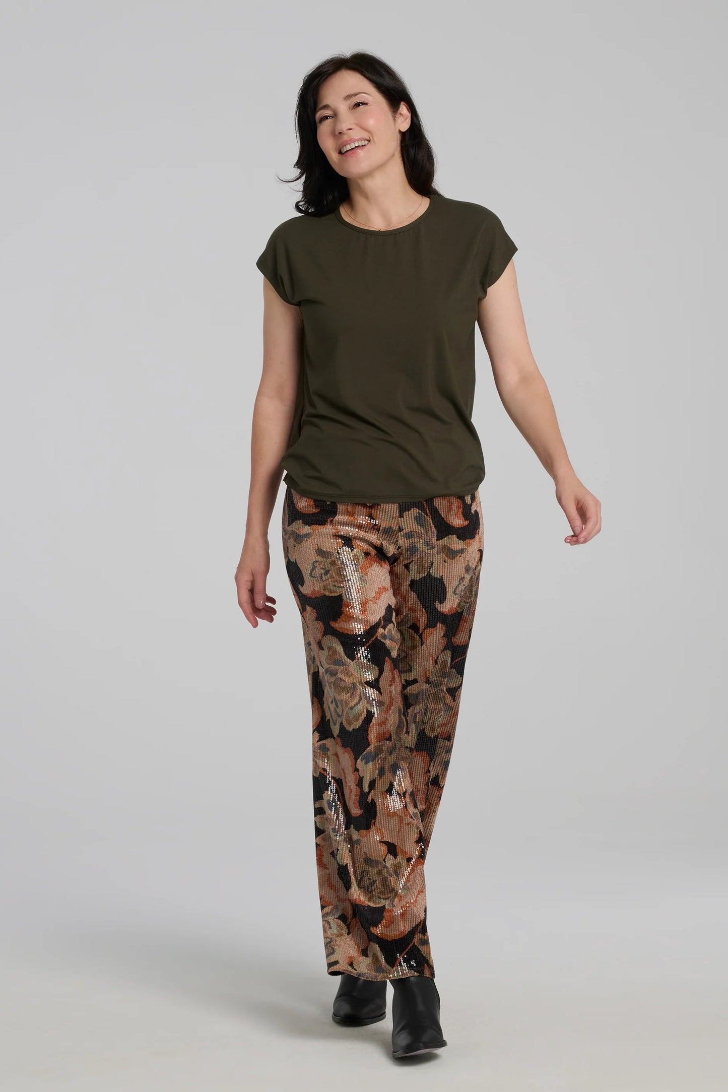 A woman wearing the Carina Pants by Kollontai, featuring floral velvet in autumn tones with clear sequins sewn right into the fabric. They have full-length straight legs and a wide pull-on waistband. She's wearing them the Eowyn Top in Fir and standing in front of a grey background. 