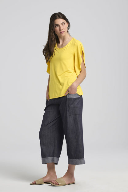 A woman wearing the Morwenna Pants from Kollontai, lightweight denim pants with wide cropped legs, an elastic waist, and contrasting trim on the large patch pockets and the cuffs. She is wearing them with the Morgana T-shirt in yellow and is standing in front of a white background. 