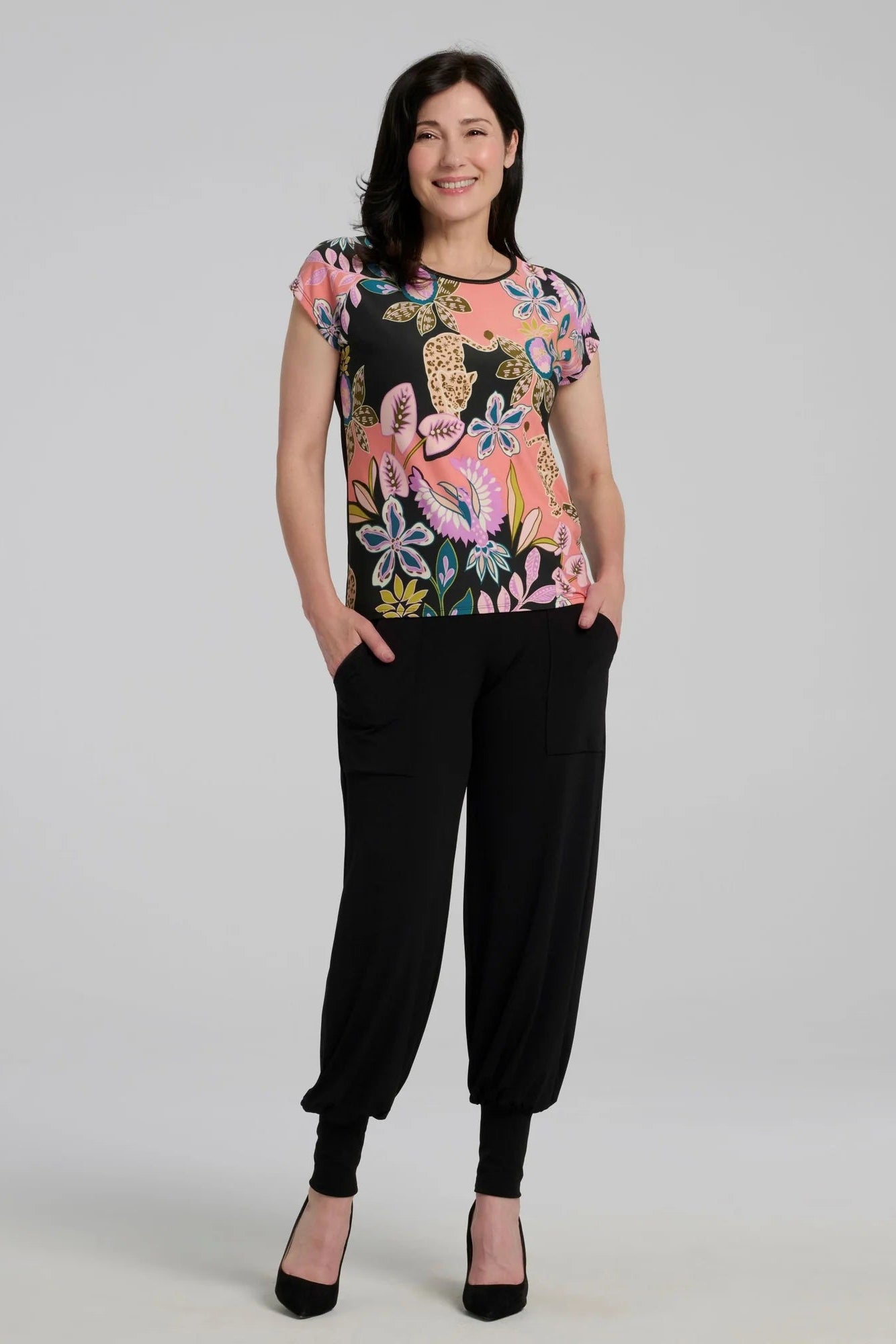 A woman wearing the Badia T-shirt by Kollontai in Bubblegum, featuring a patterned front with leopards, flowers and birds and a solid black back. It has a cap sleeves and a hi-low hem She i swearing it with black pants and standing in front of a grey background. 