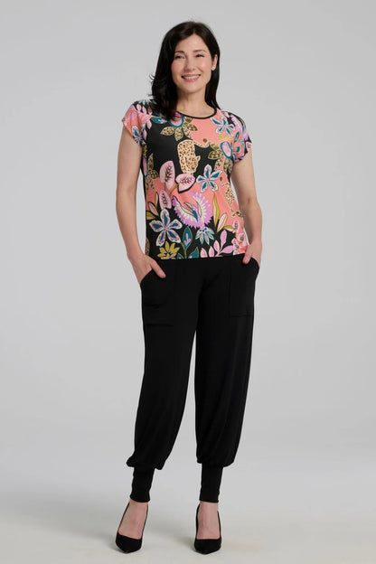 A woman wearing the Badia T-shirt by Kollontai in Bubblegum, featuring a patterned front with leopards, flowers and birds and a solid black back. It has a cap sleeves and a hi-low hem She i swearing it with black pants and standing in front of a grey background. 