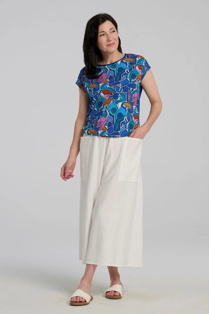Woman wearing the Hanni Top by Kollontai in Admiral, a cap sleeved relaxed top with a jewel-toned parrot pattern on the front a solid blue back. She is wearing it with white pants and standing in front of a grey background. 
