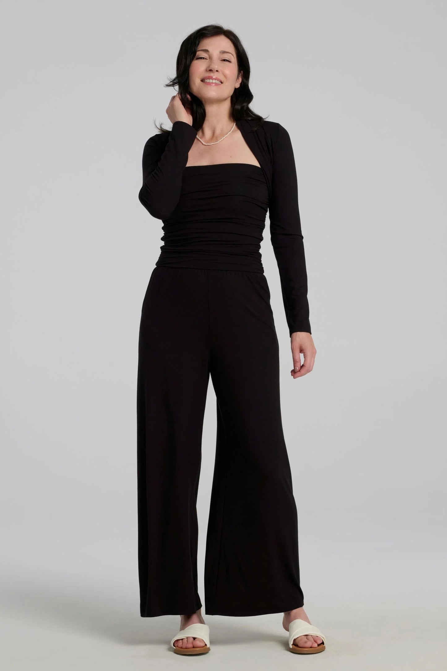 A woman wearing the black strapless Dolora Jumpsuit from Kollontai, with wide palazzo legs, with the Osimo Bolero, standing against a grey background.