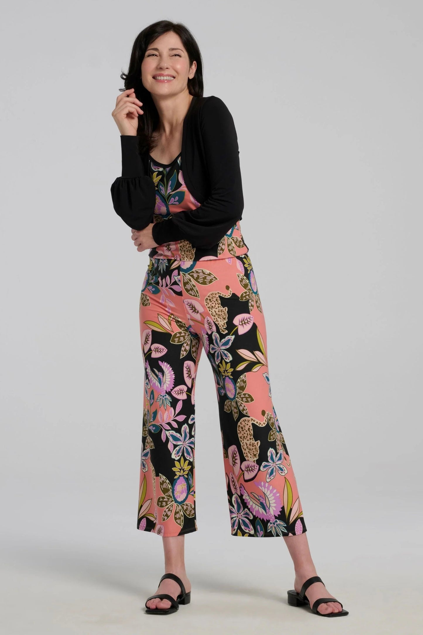 A woman wearing the Ortiga Jumpsuit by Kollontai, featuring a cheetah/floral print, spaghetti straps, an elastic waist, and 7/8 straight legs. She is wearing it with a black bolero and standing in front of a grey background.