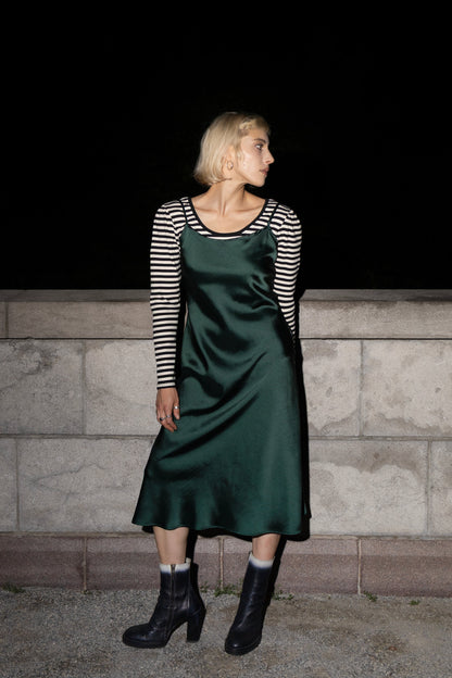 A woman wearing the Celestite Dress by Eve Lavoie in Green Satin, a slip dress with slanted spaghetti straps and an opening at the back with a tie detail. It falls to mid calf. She wearing it over a striped long sleeved top and is standing outdoors by a stone wall. 