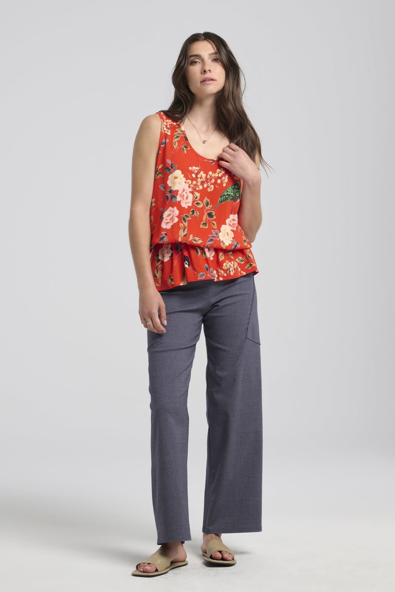 A woman wearing the Frydek Pants by Kollontai in Denim, featuring wide legs, a wide pull-on waistband, and angled patch pockets. She is wearing them with a floral tank top and standing in front of a white background. 