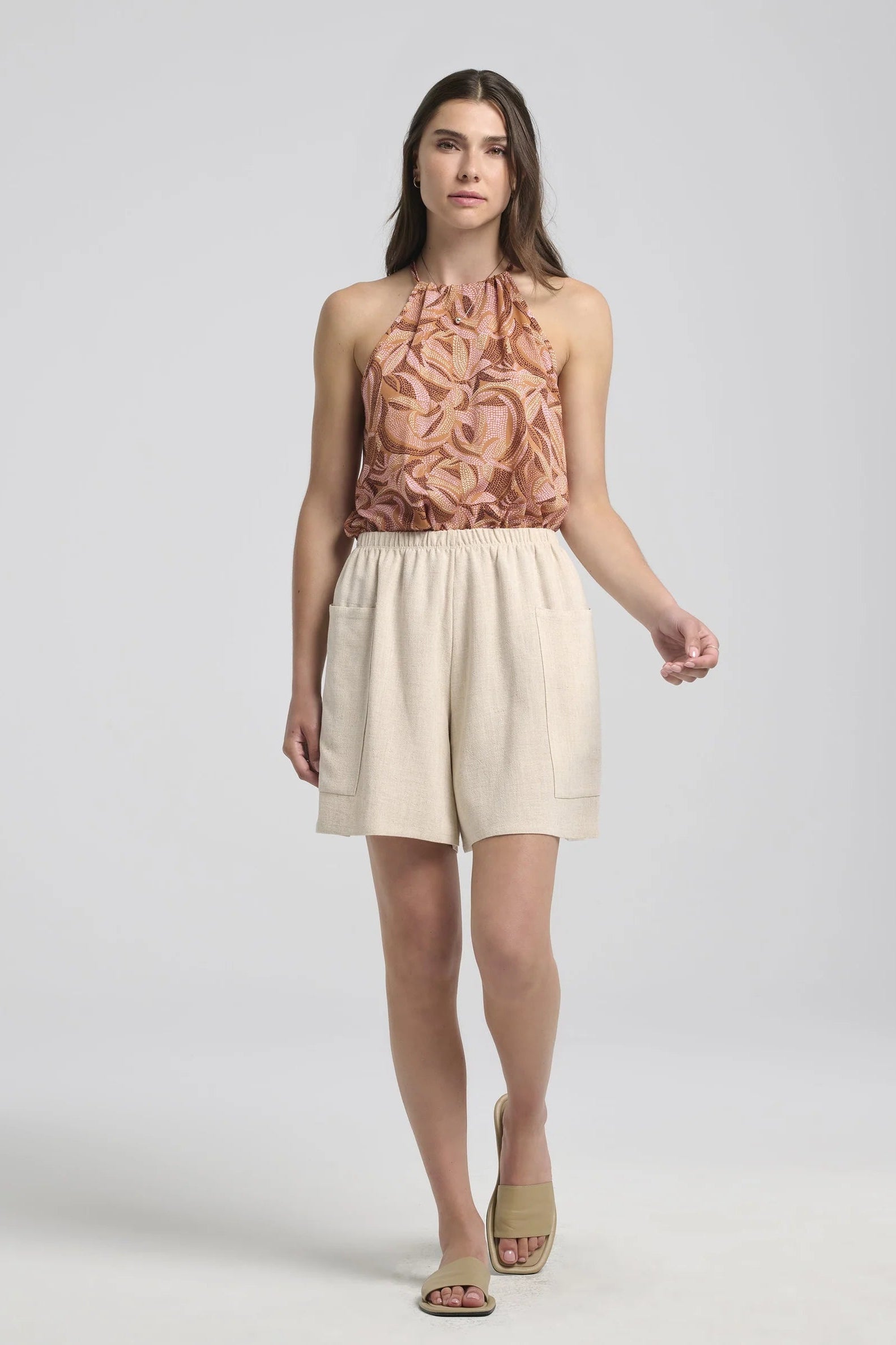 A woman wearing the Semira Camisole by Kollontai in Cinnamon print, a lightweight top with an adjustable halter neck and a loose fit. She is wearing it with a cream shorts and standing in front of a white background. 