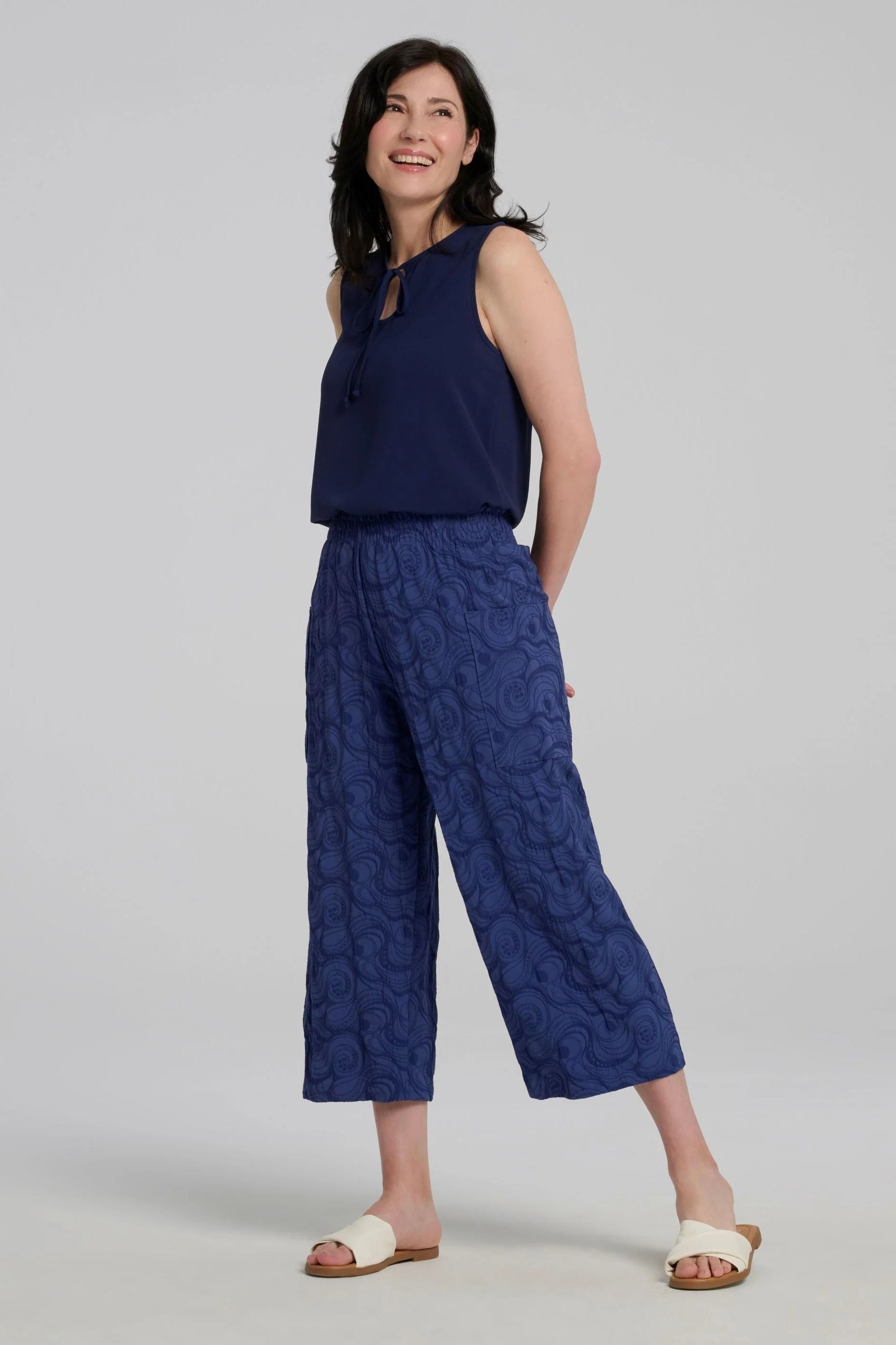 Woman wearing the Sana Pants by Kollonta in Atlantis Blue, slightly cropped straight leg pants with side patch pockets in a curlicue patterned jacquard fabric. She is wearing them with a navy sleeveless top against on a grey background.