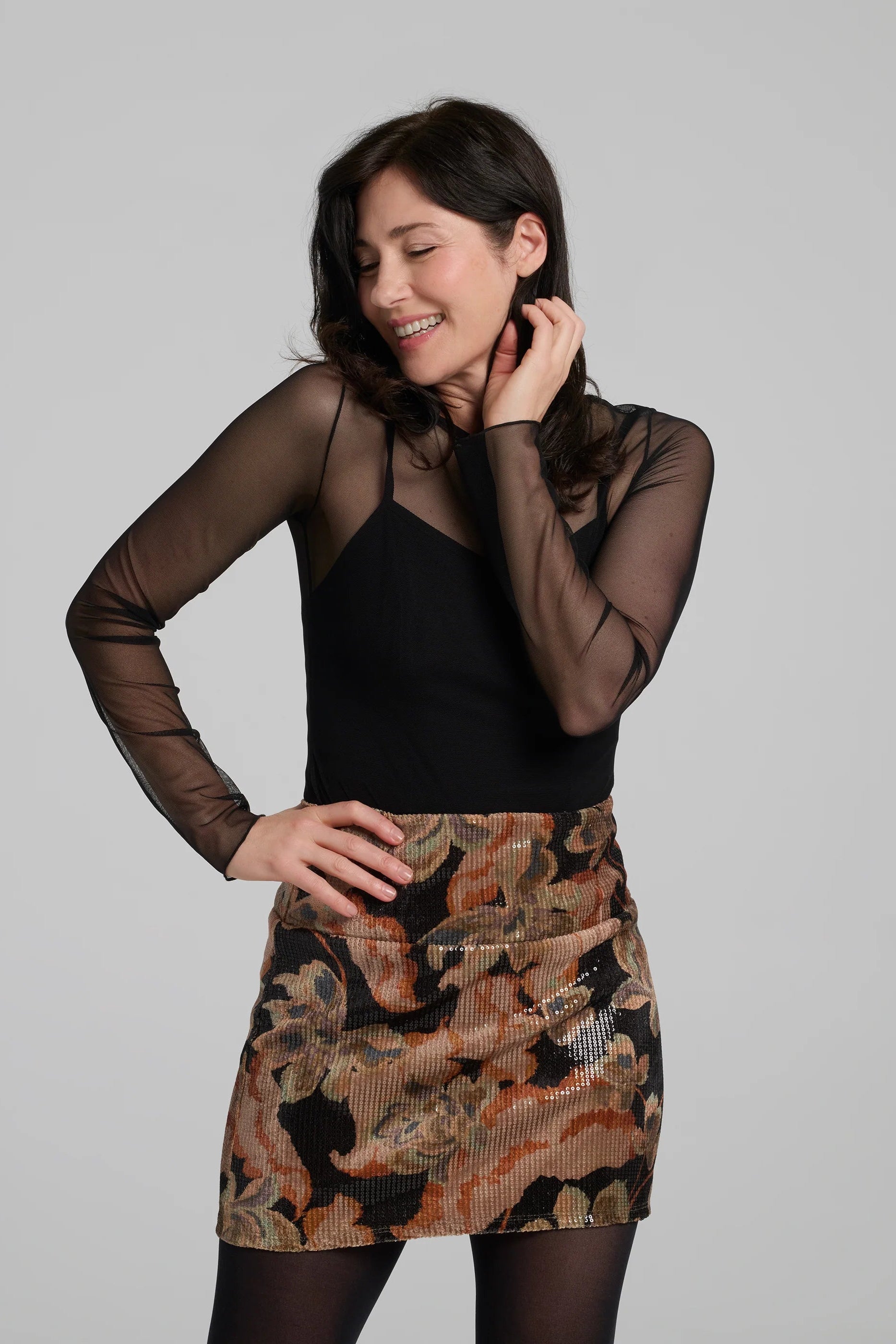 A woman wearing the Soleste Skirt by Kollontai, a floral velvet miniskirt in autumn tones with clear sequins sewn right into the fabric. It has a wide pull-on waistband. She is wearing it with a black top and standing in front of a grey background. 