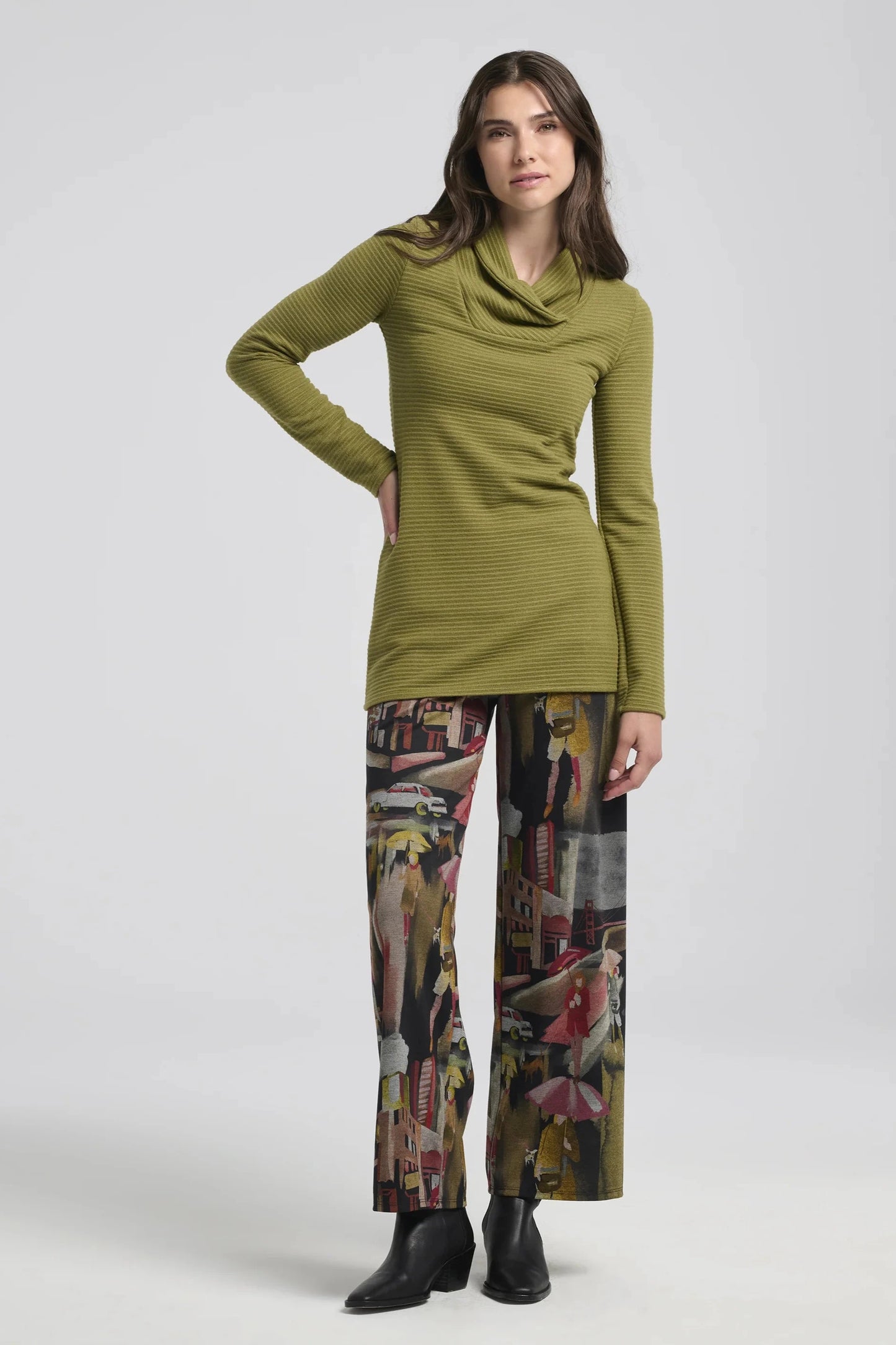 A woman wearing the Beaufort Sweater by Kollontai in Green, featuring a cross-over cowl neck, long sleeves and a ribbed knit, with patterned pants. 