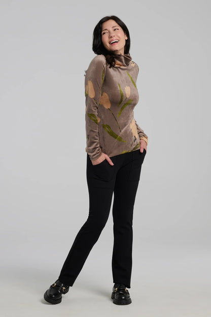 A woman wearing the Everdeen Turtleneck by Kollontai in Taupe, a velvet top with a floral print. She is wearing it with black pants and standing in front of a grey background. 