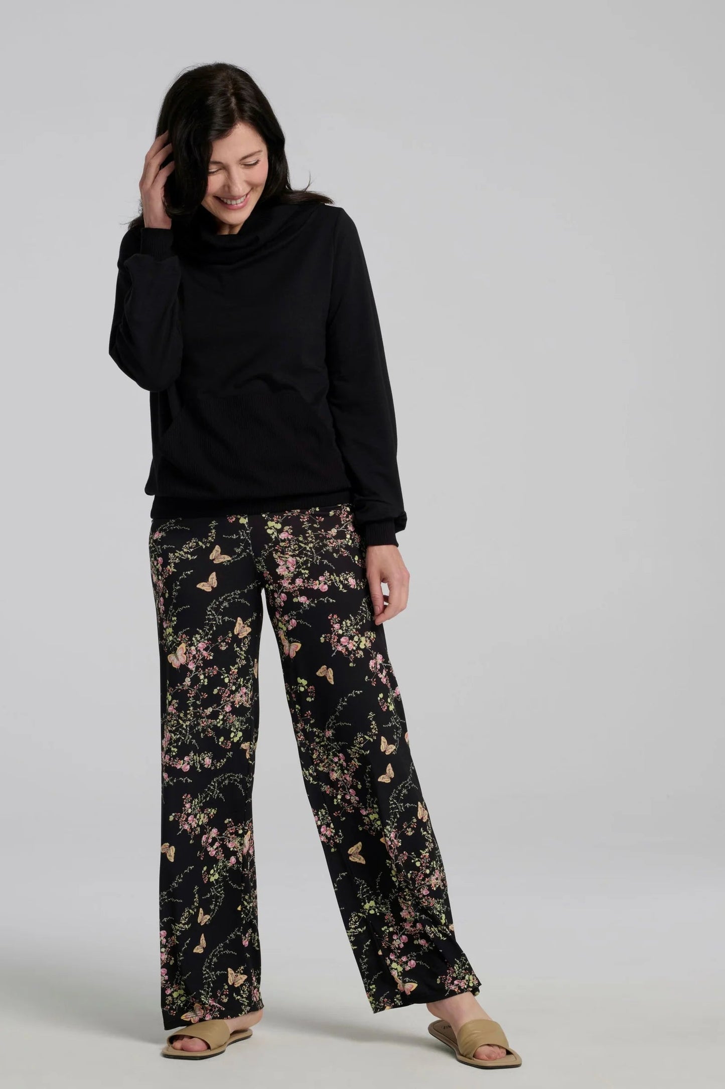 A woman wearing the Jullian Sweater by Kollontai in Black, with a cowl neck, long sleeves, and a kangaroo pocket. She is wearing it with floral pants and standing in front of a white background. 