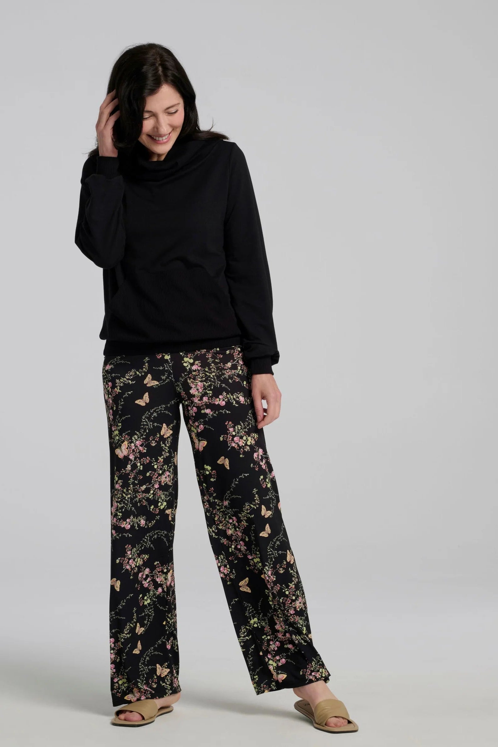 A woman wearing the Jullian Sweater by Kollontai in Black, with a cowl neck, long sleeves, and a kangaroo pocket. She is wearing it with floral pants and standing in front of a white background. 