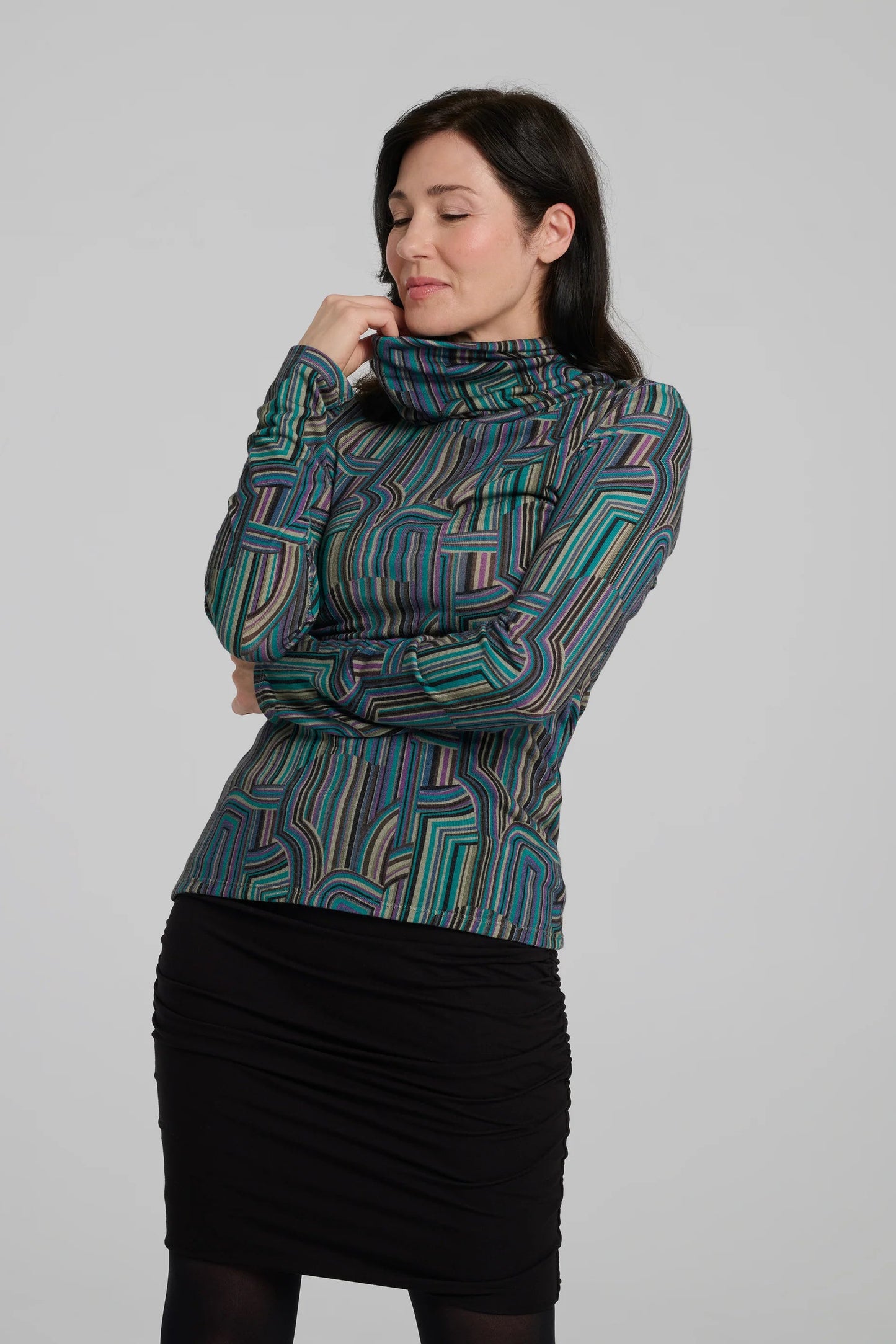 A woman wearing the Yrene Top by Kollontai in Turquoise, a geometric print turtleneck. She is wearing it with a black skirt and standing in front of a grey background. 